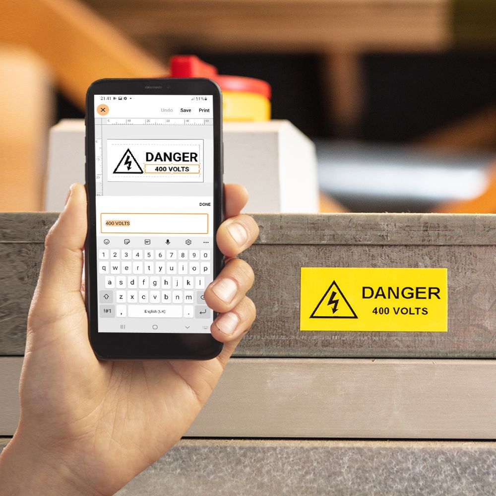 A hand holds a smartphone showcasing a design crafted with the Pro Label Tool for a Danger 400 Volts warning sign. In the background, the same cautionary message is affixed to a metal surface, reinforcing its critical importance.