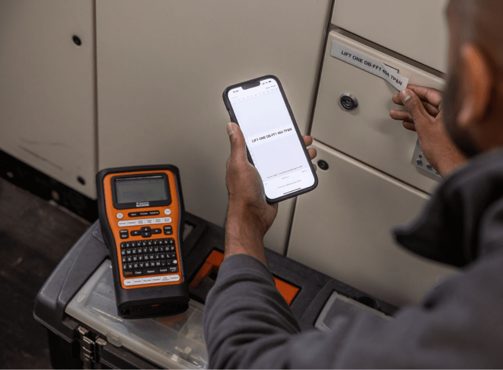 A person is using a smartphone to read instructions while holding a label printed LIFT ONE DAY AT A TIME near a panel. A nearby P-Touch EDGE label printer sits on a toolbox as the person turns a knob with their other hand, perhaps hoping for some luck in an exciting giveaway.