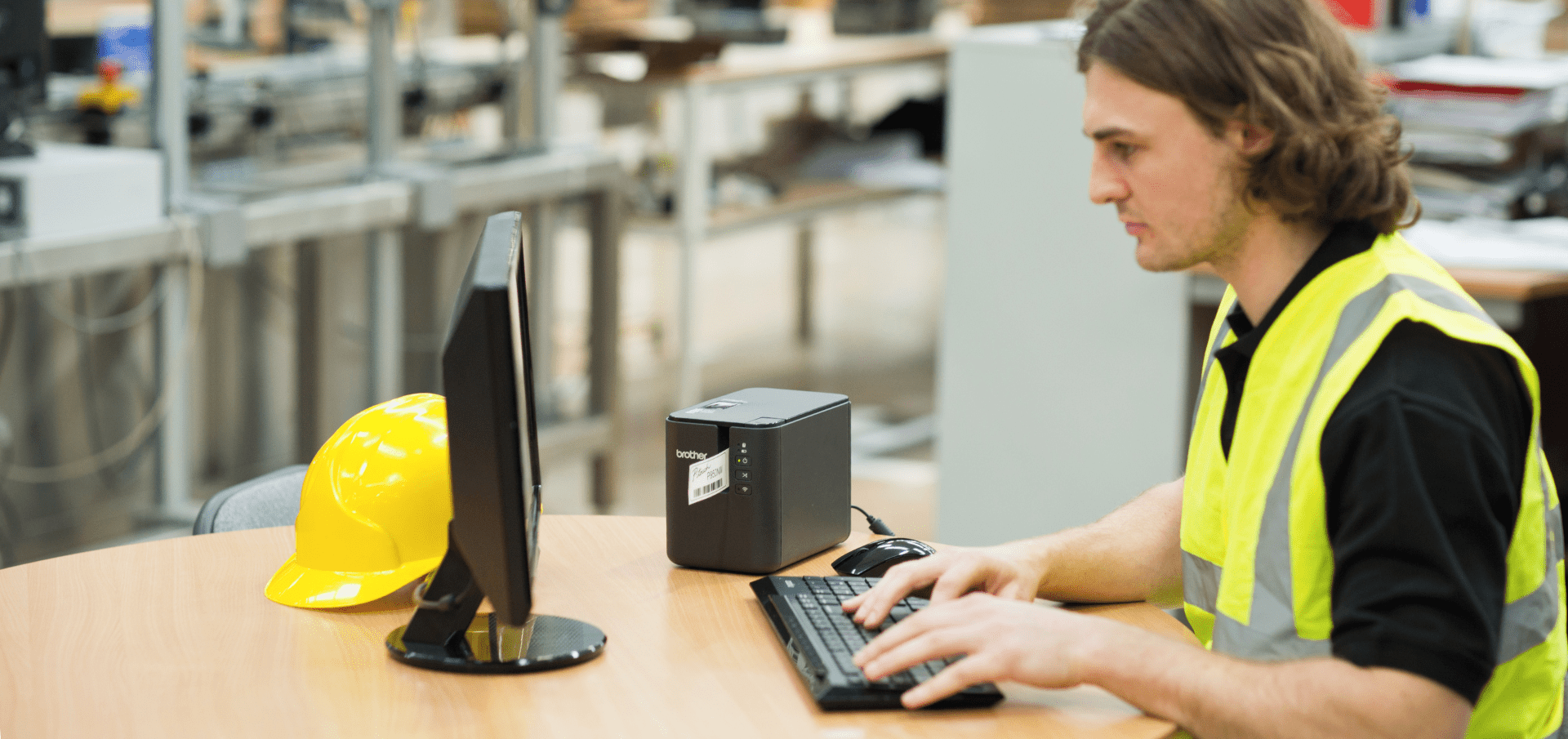 A person in a yellow safety vest sits at a desk, typing on a keyboard. A yellow hard hat and a desktop monitor are on the table. The background shows an industrial setting with machinery.