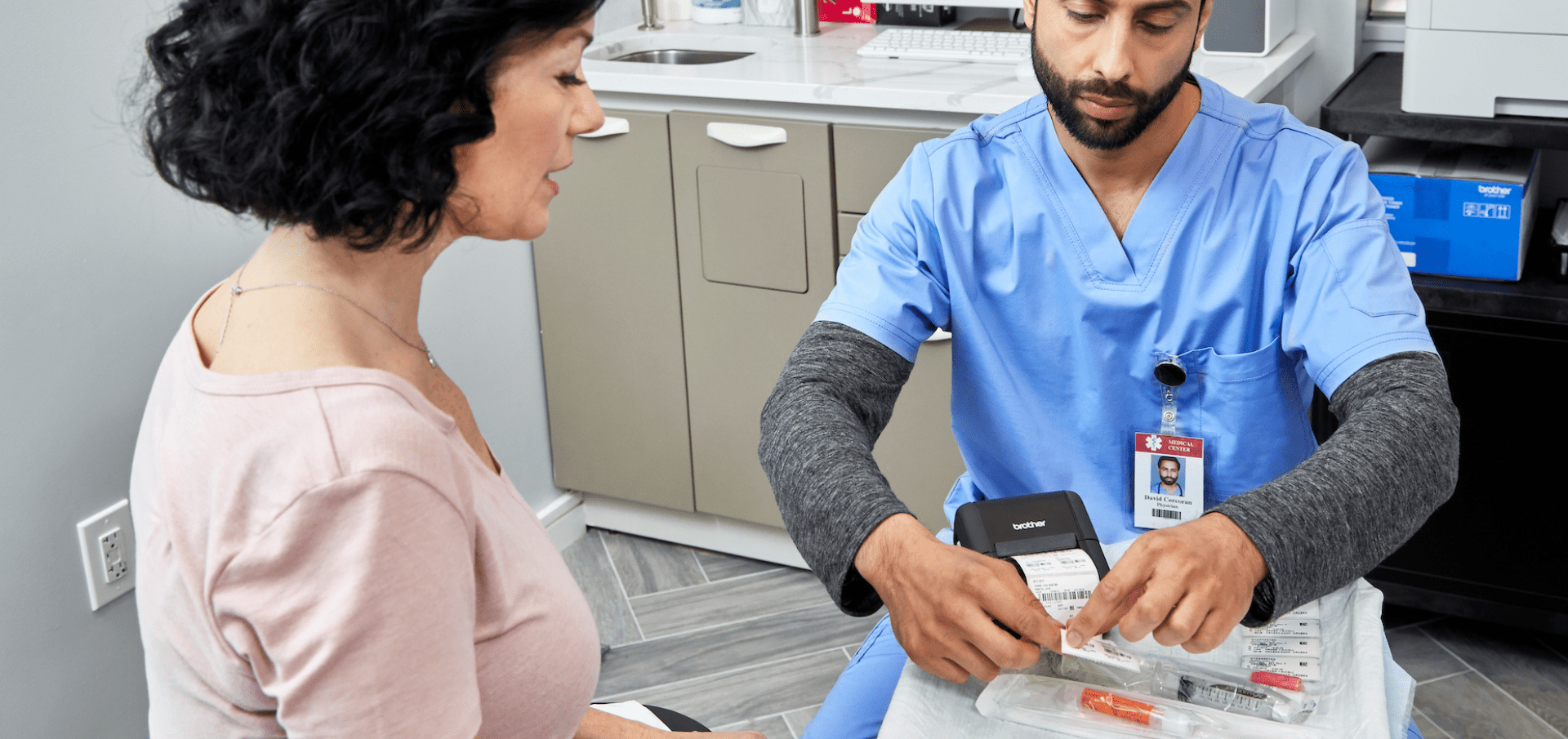 A healthcare worker in scrubs prepares an injection while talking to a seated woman in a clinic.