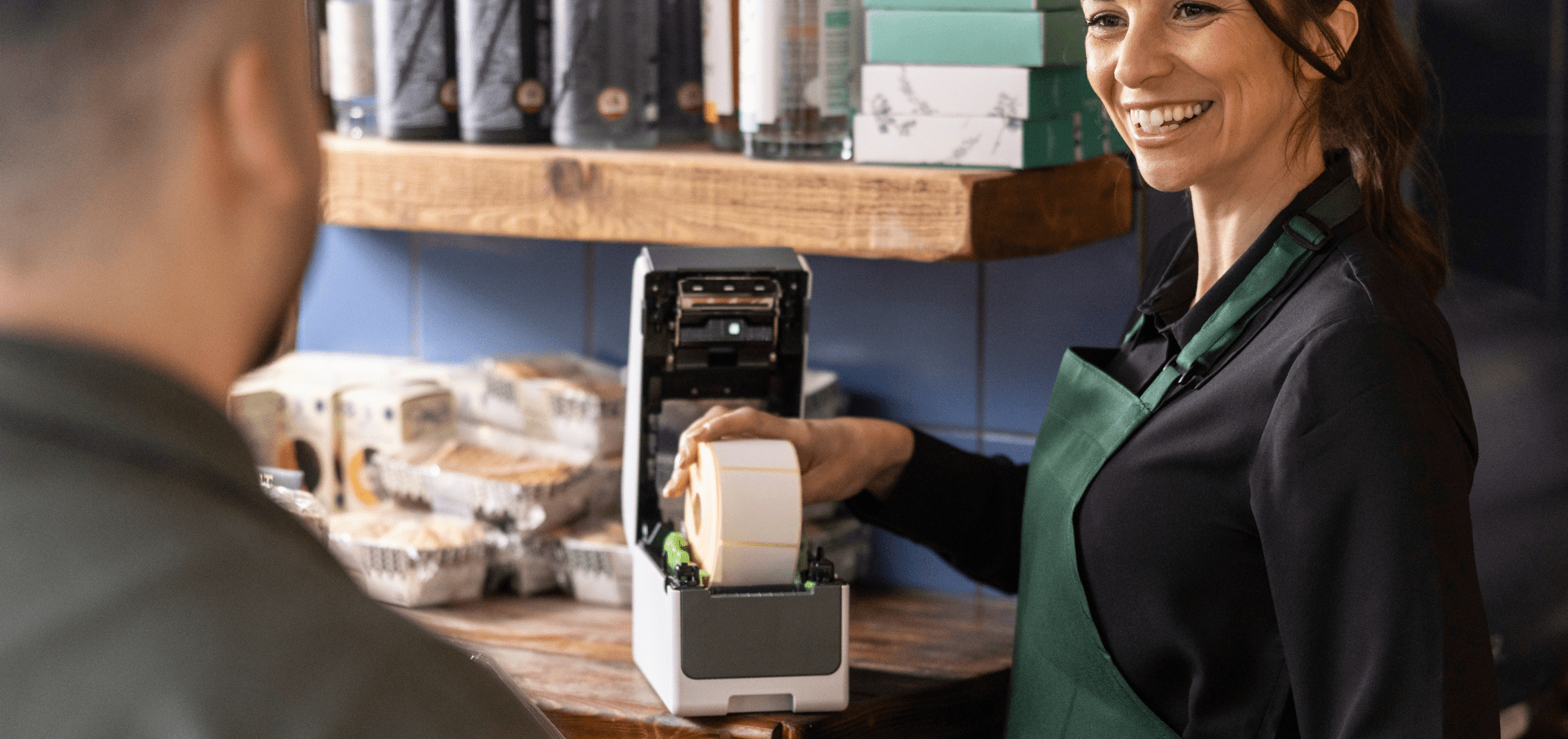 A smiling shop assistant in a green apron uses a TD 2a label printer at the counter. A customer stands in front, browsing various packaged items displayed on wooden shelves in the background.