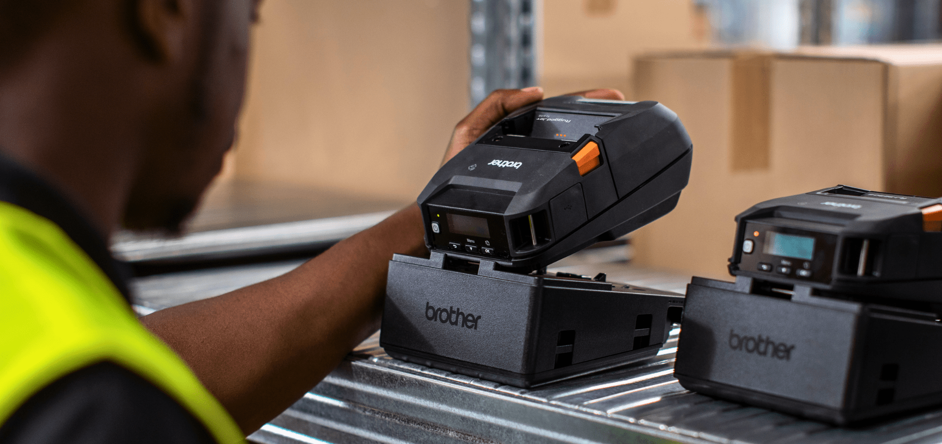 A worker places a portable Brother label printer onto a charging dock in a warehouse setting.