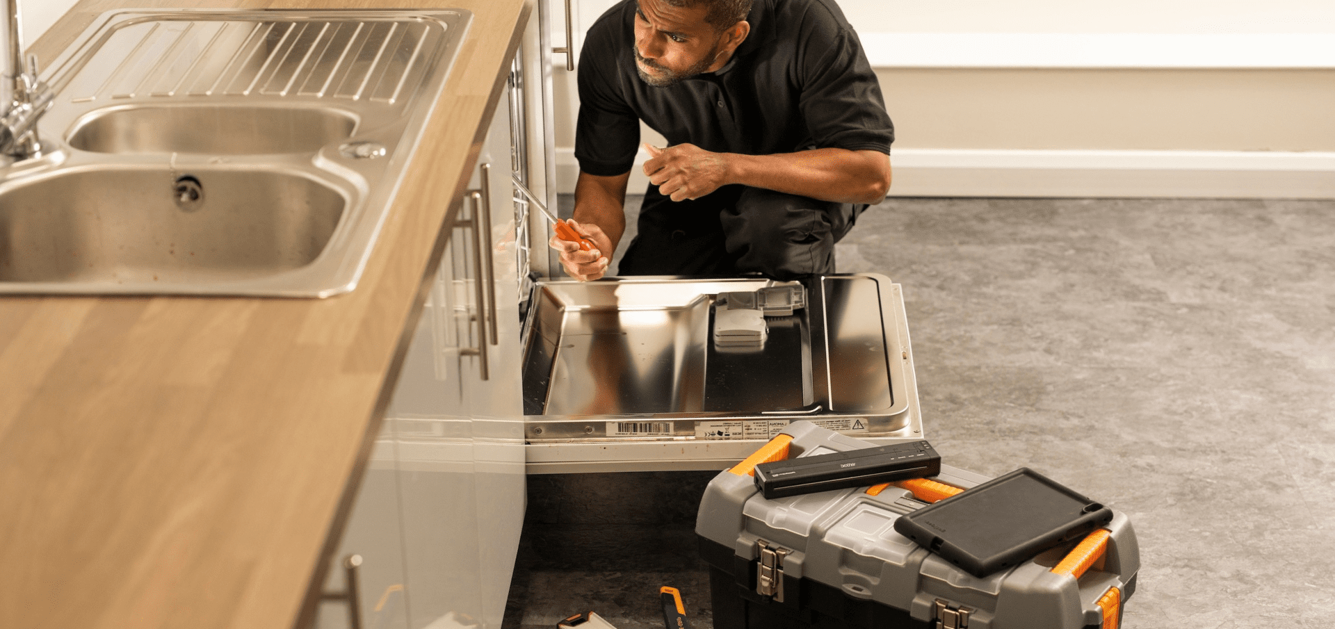 A man kneels to repair a dishwasher in a kitchen, with tools and a toolbox on the floor nearby.