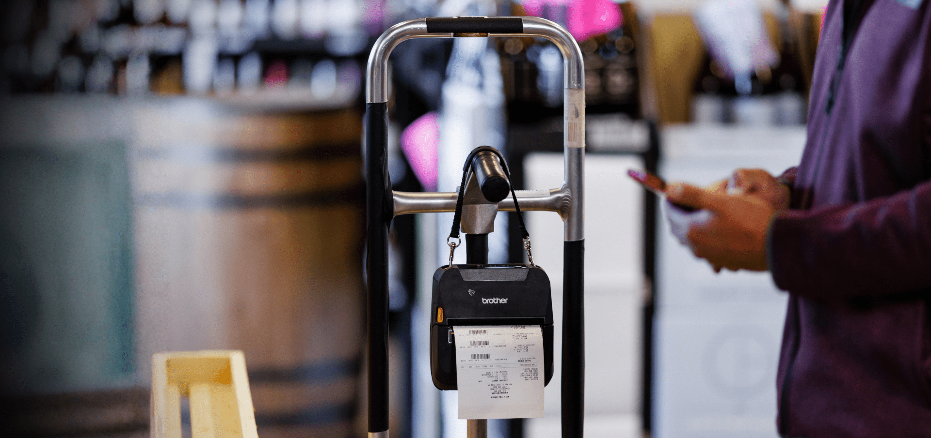 A person holds a phone near a label printer attached to a cart in a store.