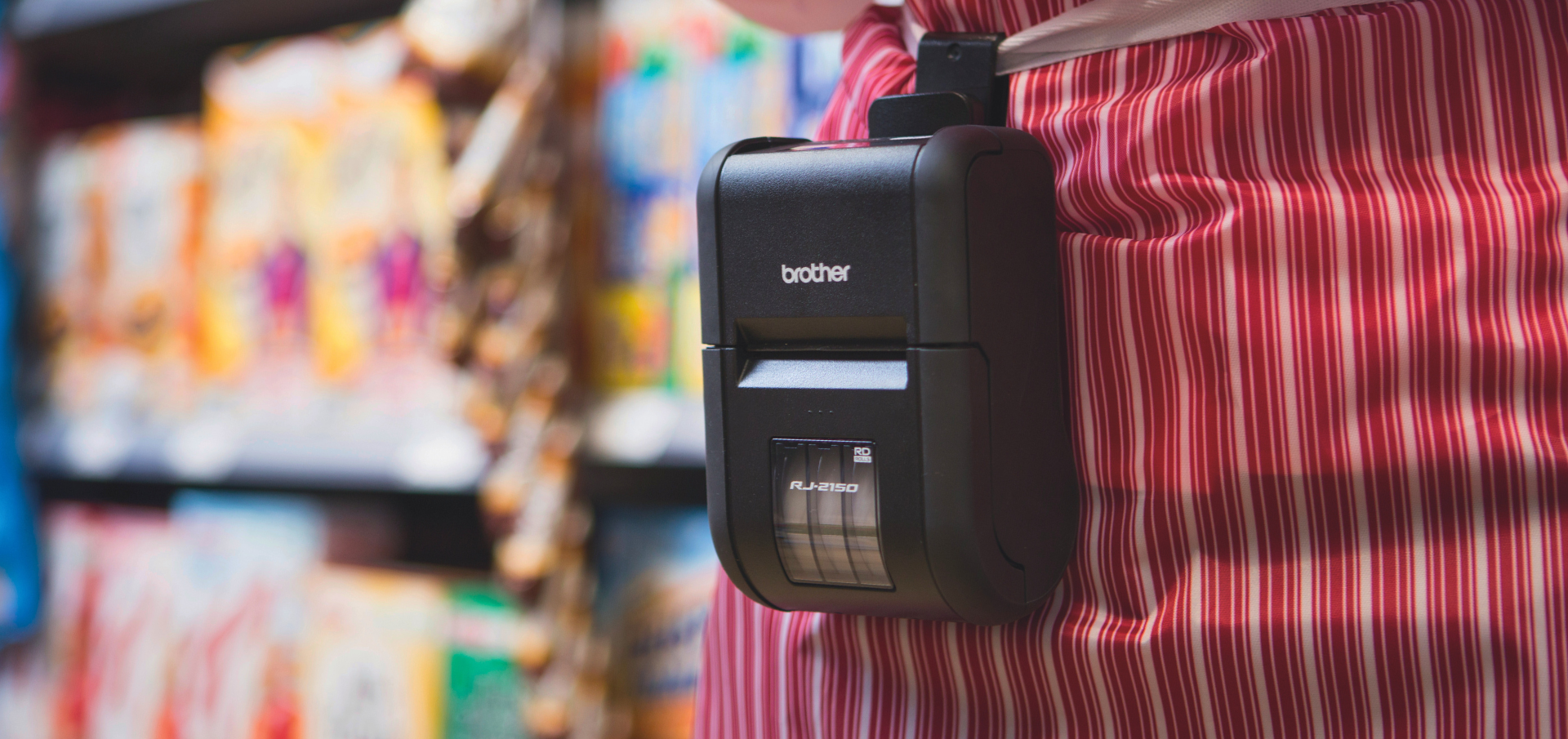 A portable Brother label printer clipped to a persons striped apron in a grocery store aisle.