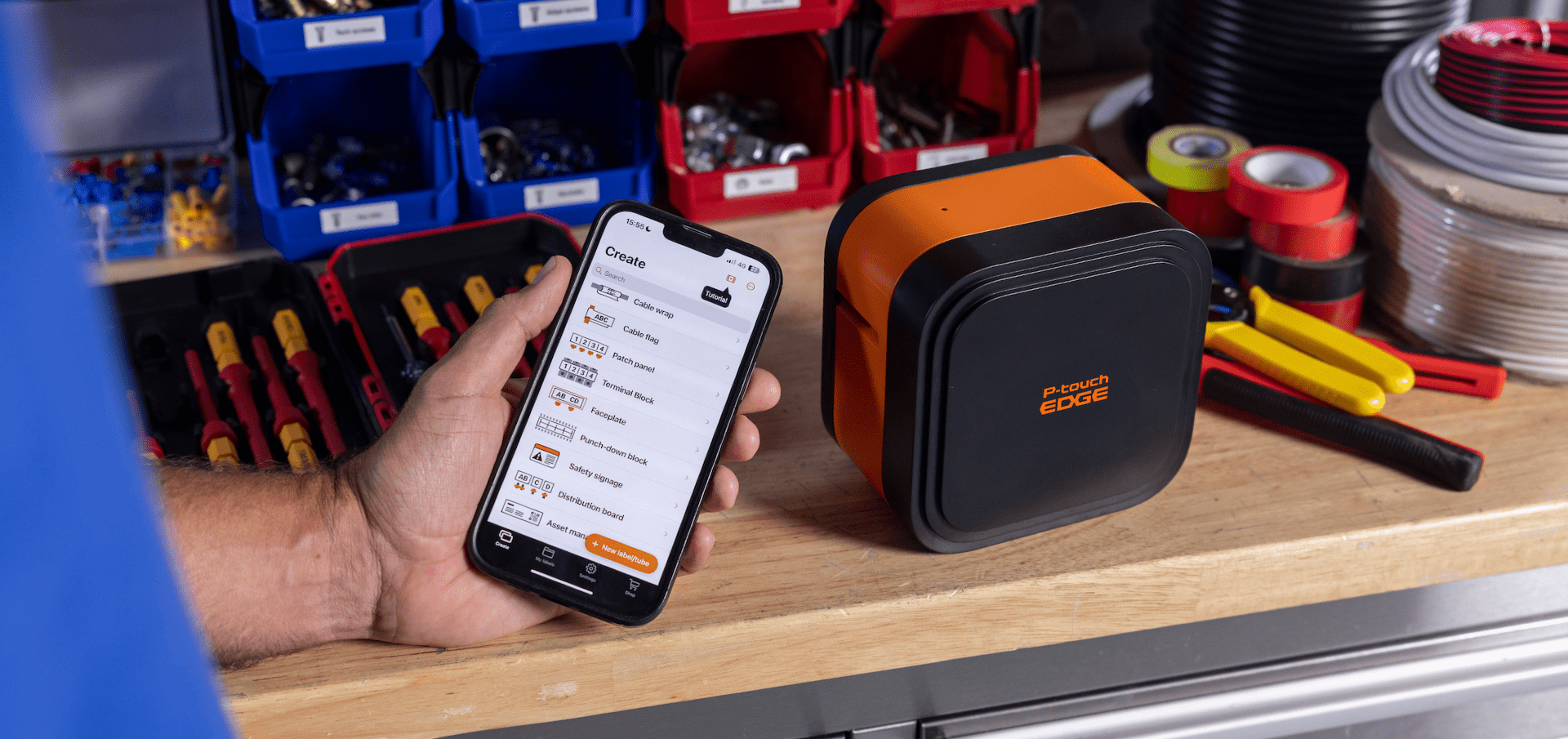 A person holds a smartphone displaying an app beside a P-touch EDGE 720BT label maker, orange and black, on a workbench.