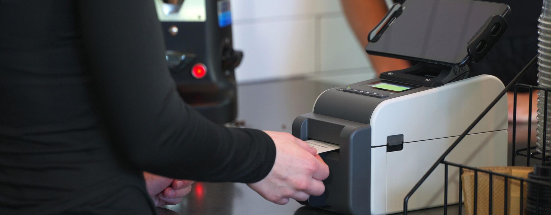 A person uses a receipt printer at the counter, seamlessly integrating linerless printing technology with a nearby touch screen device.