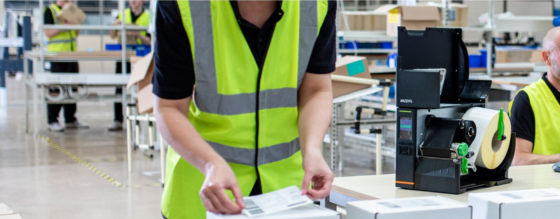 A worker in a high-visibility vest manages packages in a bustling warehouse. Nearby, a label printer with a precise printhead sits on a desk. In the background, other workers and industrial shelving highlight the efficiency of this manufacturing hub.