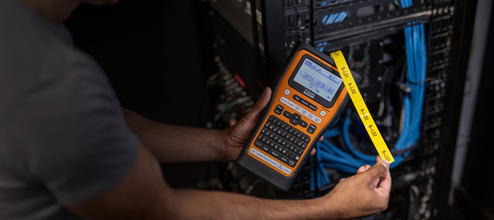 A person is holding a P-Touch EDGE label printer, carefully applying a yellow label onto a network server rack filled with blue cables. Participate in our giveaway for a chance to win your own sleek labeling tool!