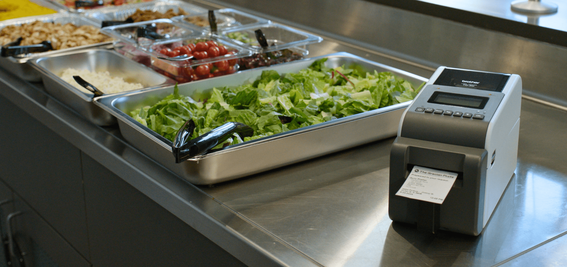 A salad bar with fresh greens and toppings next to a label printer on a stainless steel countertop.